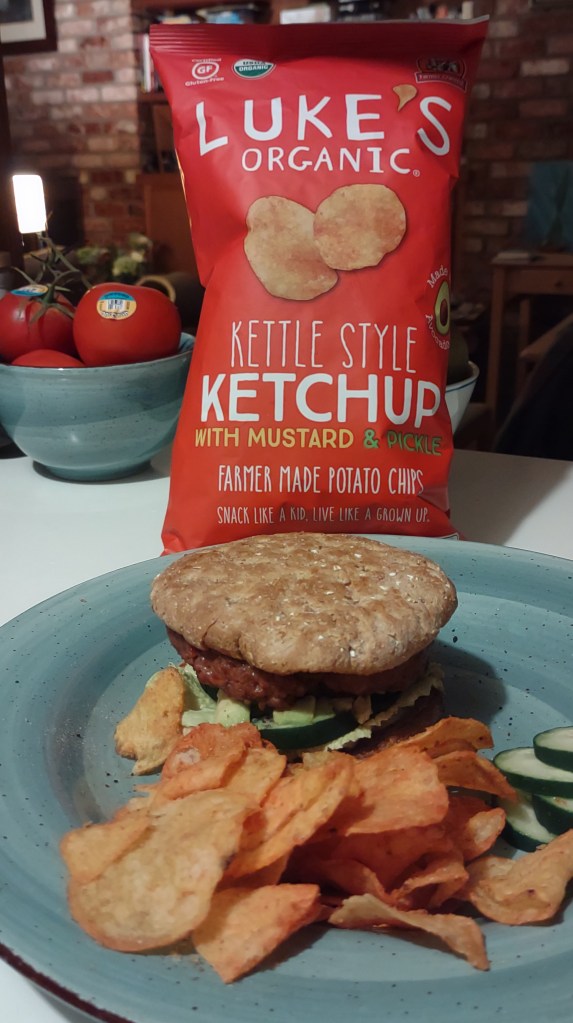 Luke’s Organic Ketchup with Mustard & Pickle chips are shown in a photo. A burger with these chips on the side are shown and behind it is a bag of the Luke’s Organic Ketchup with Mustard & Pickle chips. Luke’s Organic chips sustainability review