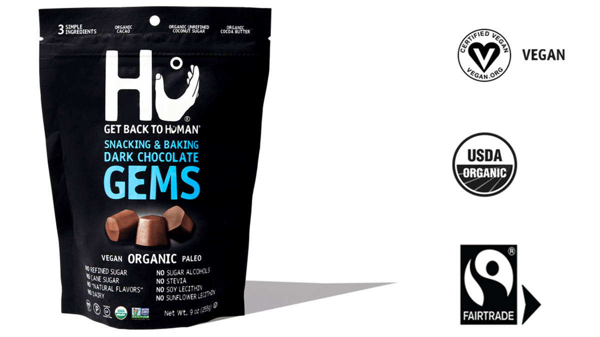 A bag of Hu Dark Chocolate Gems is pictured next to three logos indicating key certifications: vegan, organic, and fair trade. 
