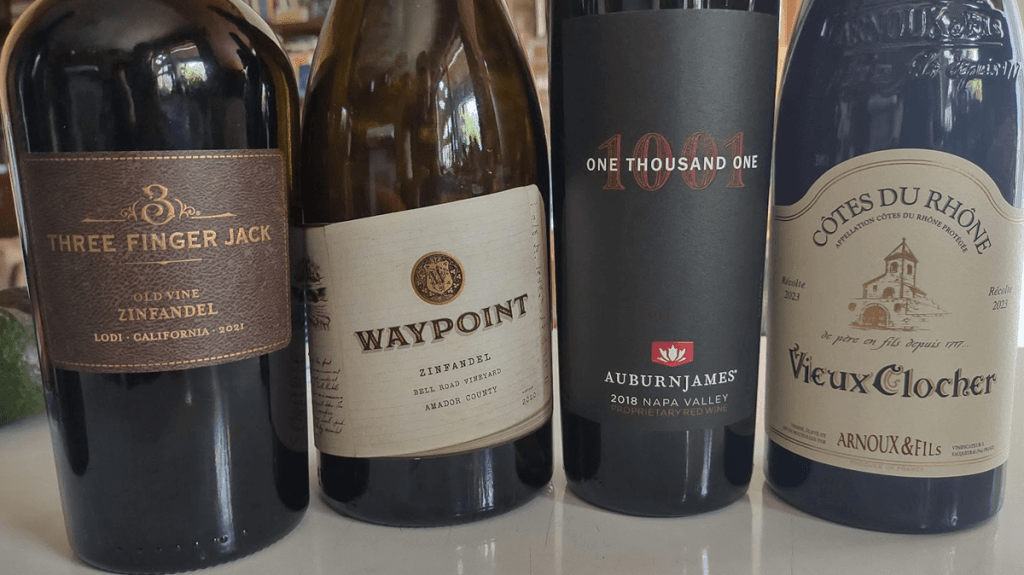 Wines to try during the Grocery Outlet spring 2025 sale. Four bottles of red wine are pictured. As described in the legend, they are: Three Finger Jack old vine zinfandel, Lodi, California ($7); Waypoint zinfandel, Amador County, California ($8); Auburn James 1001 red blend, Napa Valley, California ($15); Vieux Clocher Côtes du Rhône, France ($8). All wines will be 20% off the prices listed above, during the sale.