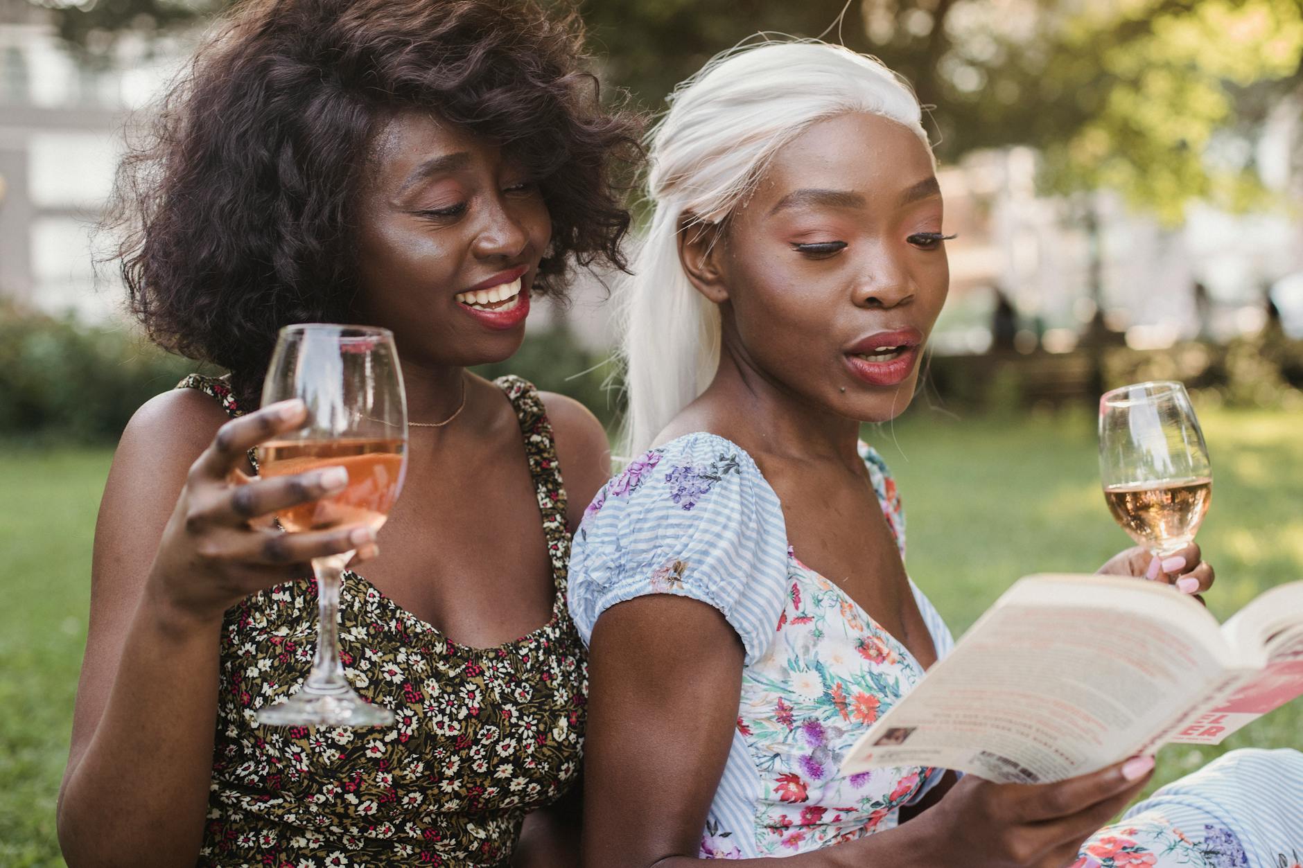 Two women sit in a park drinking wine while one of them reads from a book.